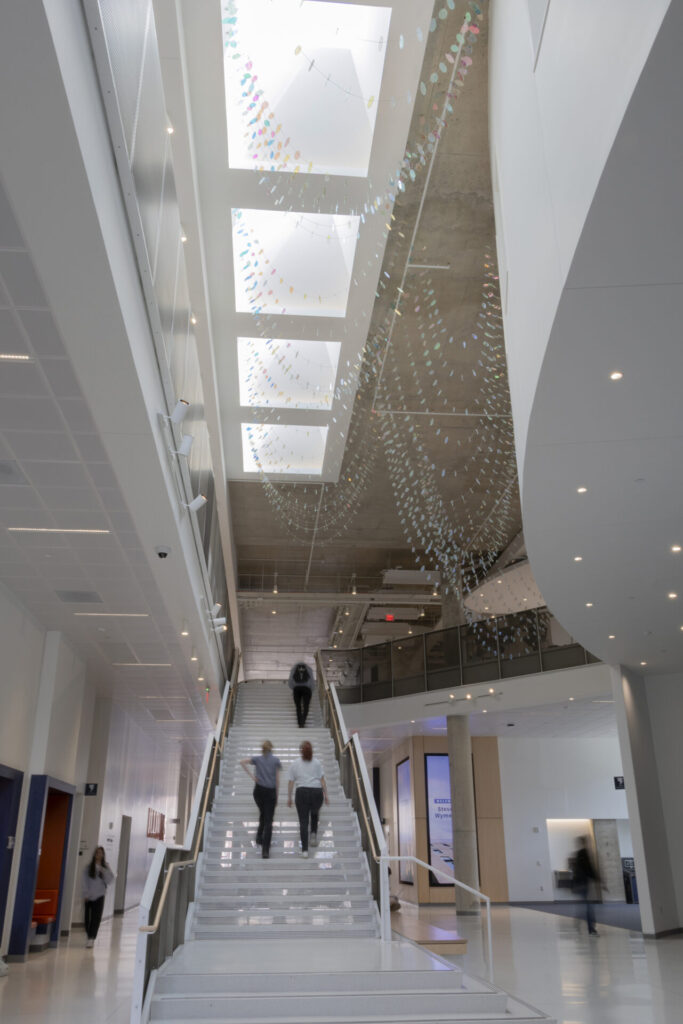 Interior view of the Wymer Hall atrium showing students on the grand stairway beneath suspended glass sculpture elements descending from five skylights