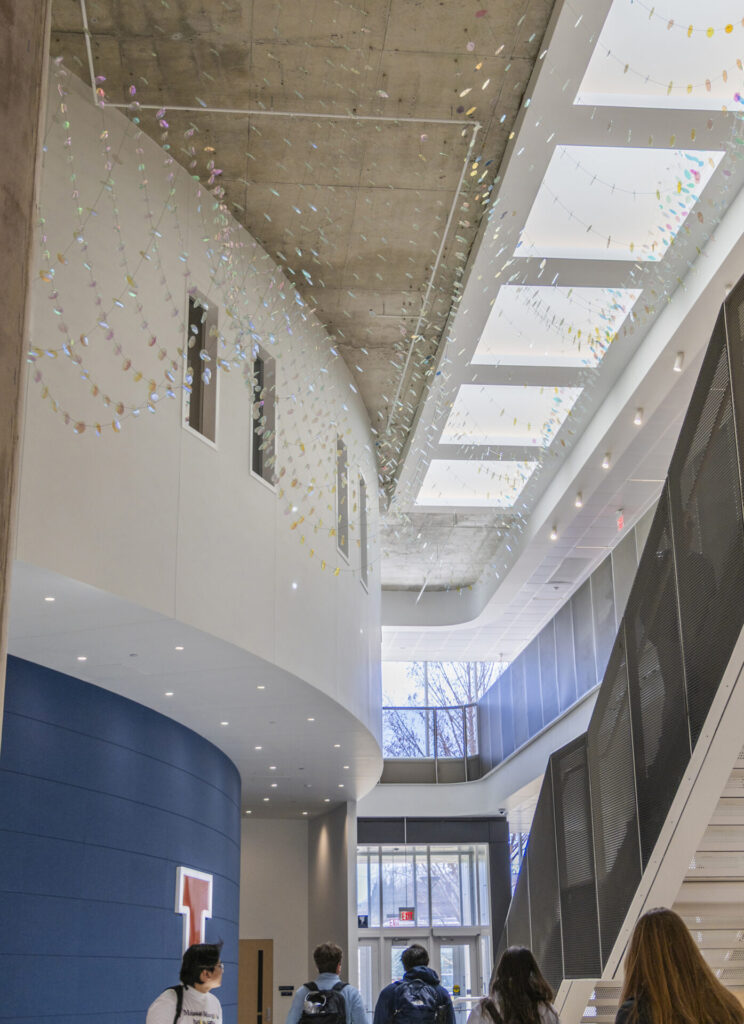 Looking up to the glass suspended sculpture on ceiling -viewed from center of the long atrium space. 5 students are walking towards the north entrance
