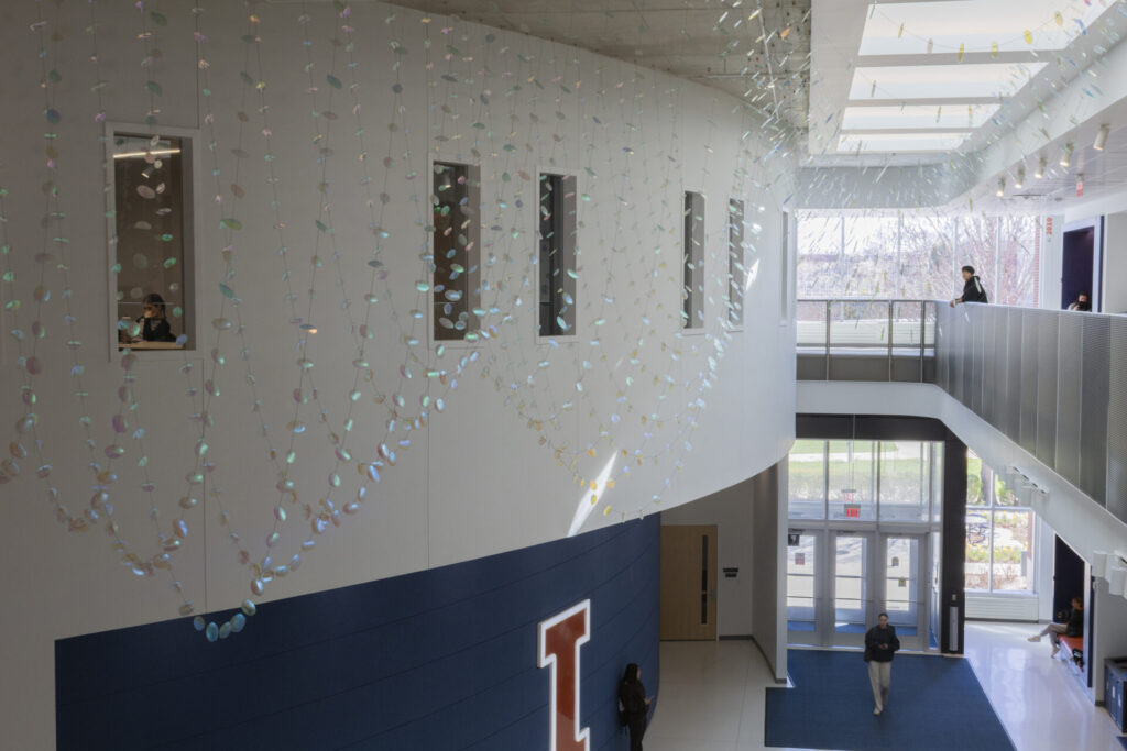 Sunlight entering from the skylight is visible on the curved wall. Glass suspended sculpture with 2,000 dichroic glass flower petals and leaves inside Steven Wymer Hall, University of Illinois Urbana-Champaign