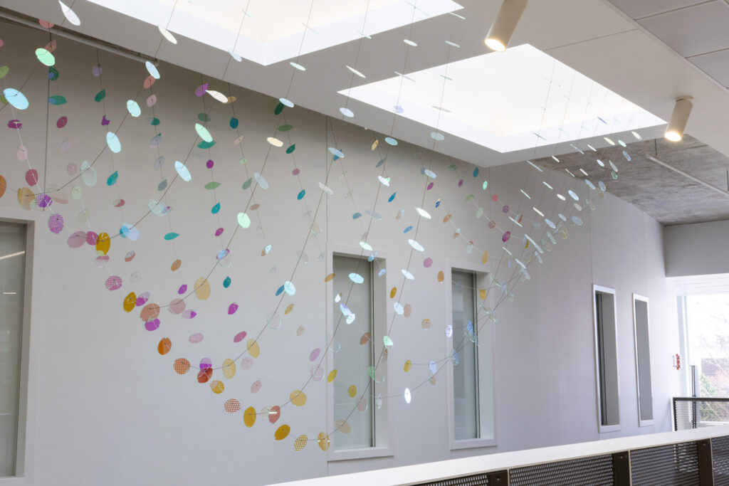 Close-up interior view of suspended dichroic glass petals and leaves at the south end of the Wymer Hall sculpture installation near a curved wall and railing