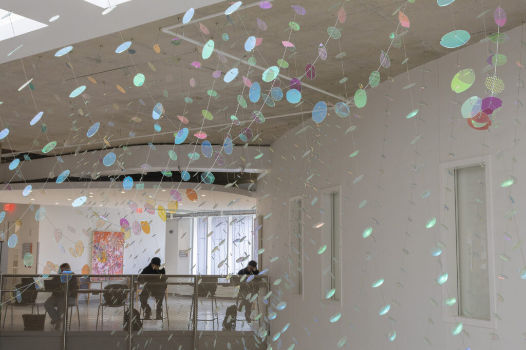 View from the second floor railing in Wymer Hall at the University of Illinois Urbana-Champaign, showing a suspended dichroic glass sculpture with close-up floating petals in the foreground and additional iridescent slumped glass forms extending toward a curved wall and student study tables.